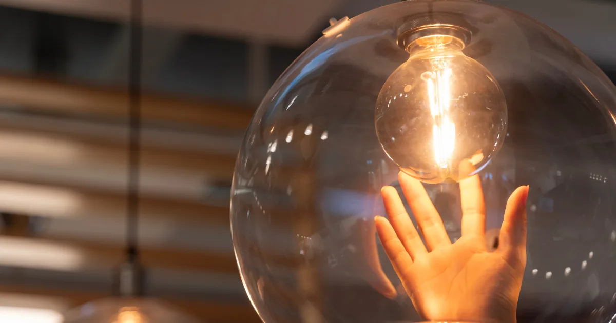 ガラスの球体照明の中で光る電球に手を伸ばすイメージ、電気代をイメージした写真